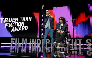 A white man with brown curly chair sits in a wheelchair in front of a microphone on a colorful stage.